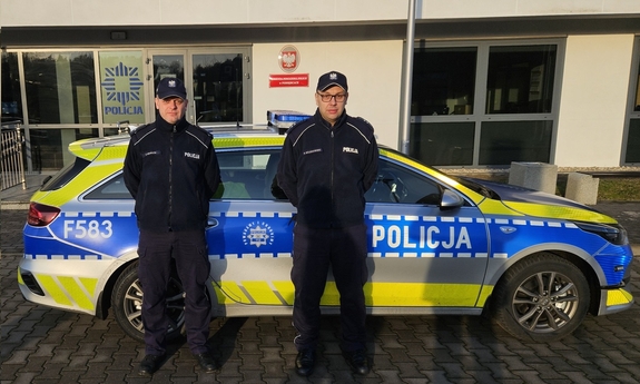 Dwaj policjanci stoją przy radiowozie policyjnym W tle budynek komendy Policji.