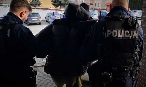 Dwaj umundurowani policjanci prowadzą zatrzymanego mężczyznę.