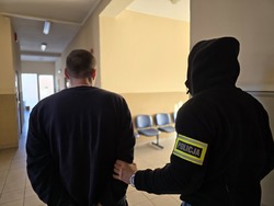 Nieumundurowany policjant w opasce na ręce z napisem Policja, prowadzi zatrzymanego. Widok z tyłu.