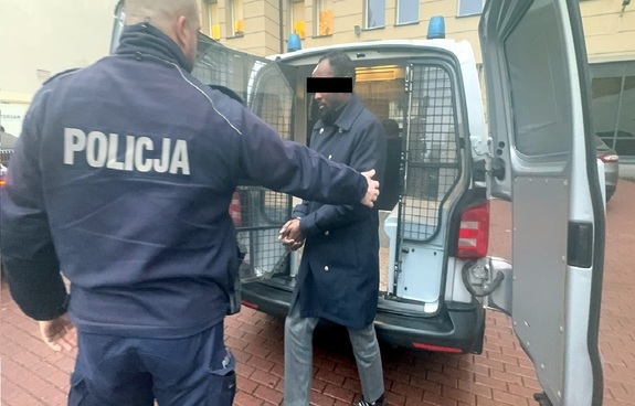 Umundurowany policjant z zatrzymanym mężczyzną przed radiowozem.