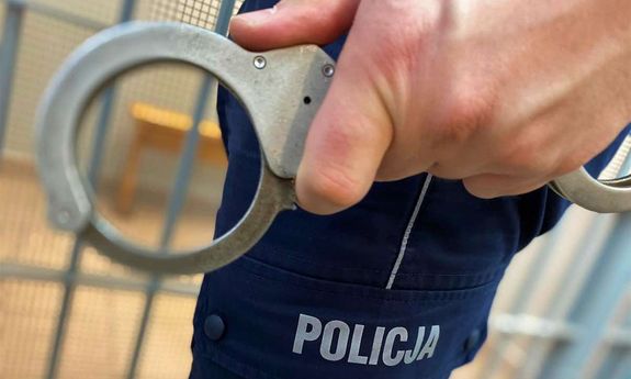 Widoczna część dłoni, w której trzymane są kajdanki, oraz nogawka spodni, na których znajduje się napis: Policja.