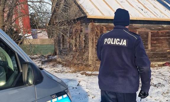 Dzielnicowy kontroluje miejsce, gdzie może przebywać osoba zagrożona wychłodzeniem. W głębi drewniany dom.