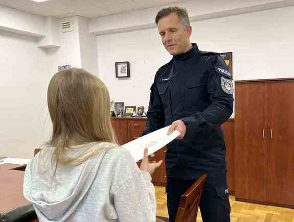 Policjant - komendant, wręcza dziewczynce podziękowania.