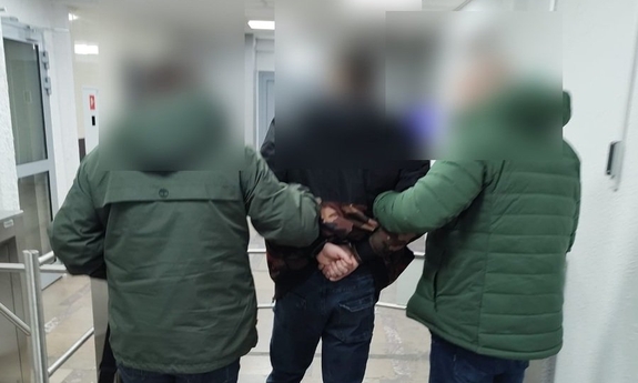 Policjanci kryminalni stojący tyłem oraz osoba zatrzymana w kajdankach.