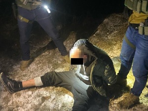 Zatrzymany mężczyzna skuty w kajdanki siedzi na ziemi, obok niego stoją dwaj funkcjonariusze.