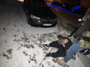 Zatrzymany mężczyzna skuty w kajdanki siedzi na ziemi, obok niego stoją funkcjonariusze. Za nim stoi samochód w kolorze czarnym.