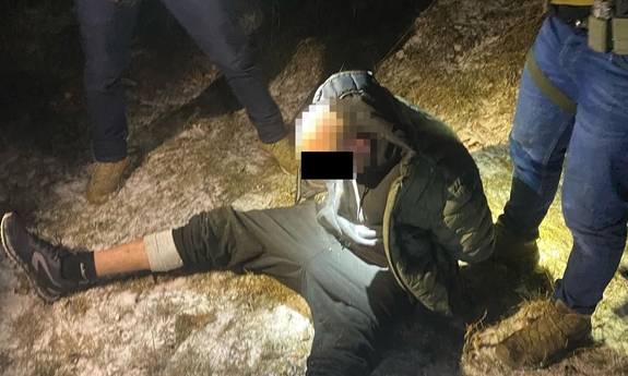 Zatrzymany mężczyzna skuty w kajdanki siedzi na ziemi, obok niego stoją dwaj funkcjonariusze.