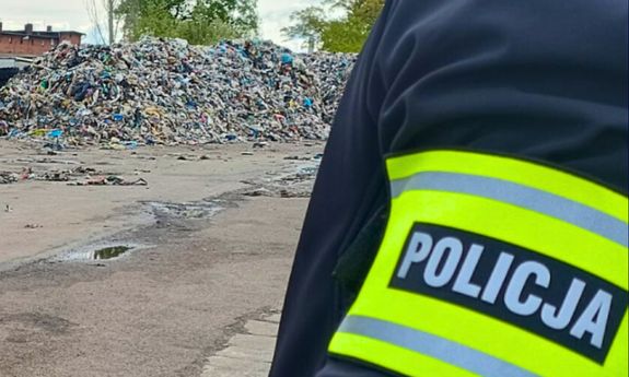 Na pierwszym planie, z prawej strony widoczny fragment ramienia policjanta, na którym znajduje się opaska z napisem Policja. W tle widać wysypisko śmieci.