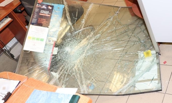 Leżąca na posadzce popękana szyba pancerna, w tle fotel i fragment mebli.