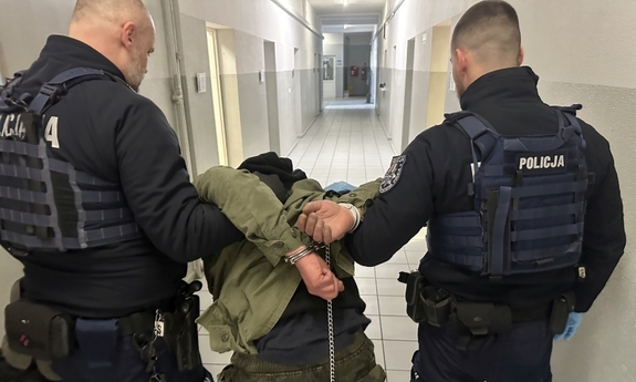 Dwaj funkcjonariusze prowadzą zatrzymanego zakutego w kajdanki.