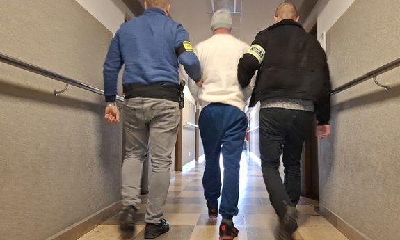 Dwaj nieumundurowani policjanci prowadzą zatrzymanego korytarzem - widok z tyłu.