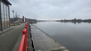 Betonowe nabrzeże, rzeka i budynki nieopodal. W tle widać stojących na nabrzeżu wędkarzy.