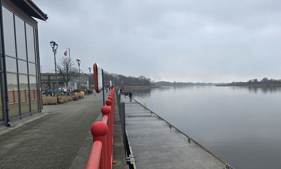 Betonowe nabrzeże, rzeka i budynki nieopodal. W tle widać stojących na nabrzeżu wędkarzy.