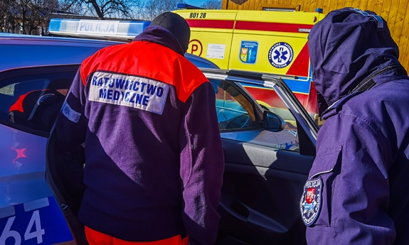 Ratownik medyczny i umundurowany policjant stoją przy radiowozie. W tle karetka pogotowia.