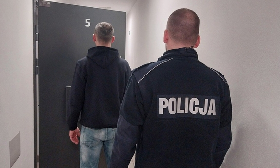 Policjant i zatrzymany mężczyzna w pomieszczeniu stojący tyłem.