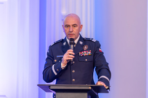 Zastępca Komendanta Głównego Policji, Rafał Kochańczyk ubrany w granatowy mundur, trzyma w dłoni mikrofon, stoi przy podium i wygłasza przemówienie.