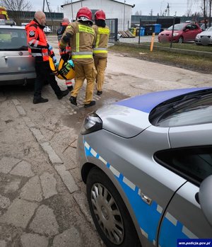 Funkcjonariusze straży i sanitariusze niosą na noszach osobę. Z tyłu widać fragment radiowozu, z przodu cywilne auto.