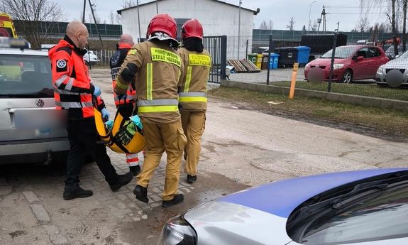 Funkcjonariusze straży i sanitariusze niosą na noszach osobę. Z tyłu widać fragment radiowozu, z przodu cywilne auto.