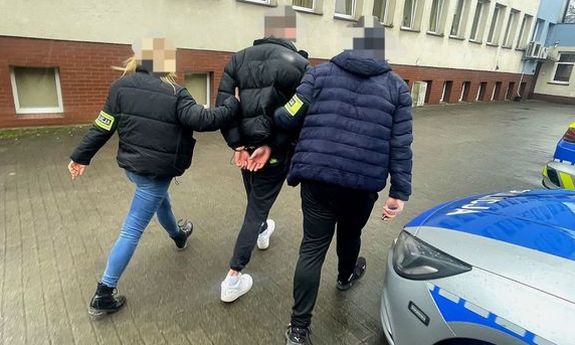 Dwoje nieumundurowanych policjantów prowadzi zatrzymanego. Z boku widać fragmenty dwóch radiowozów. Widok z tyłu.