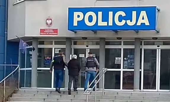 Dwaj funkcjonariusze wprowadzają do budynku komendy Policji po schodach zatrzymanego mężczyznę w kajdankach.