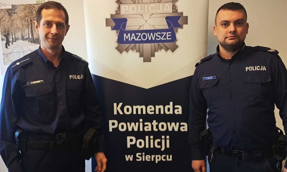 Dwóch umundurowanych policjantów pozuje do zdjęcia obok baneru Komendy Powiatowej Policji w Sierpcu.