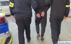 Dwaj funkcjonariusze prowadzą mężczyznę zakutego w kajdanki.