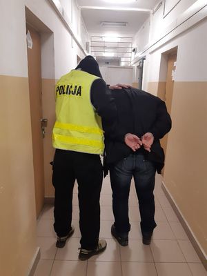 Zatrzymany mężczyzna zakuty w kajdanki prowadzony przez policjanta korytarzem.