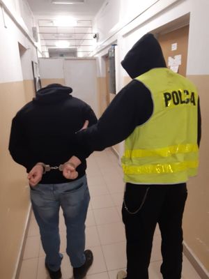 Zatrzymany mężczyzna zakuty w kajdanki prowadzony przez policjanta korytarzem.
