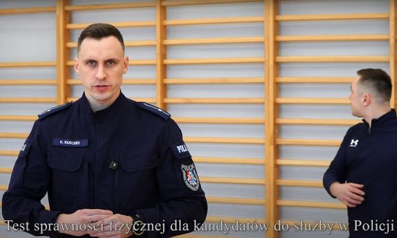policjant stoi przodem do kamery, za nim ćwiczy przy drabinkach mężczyzna.