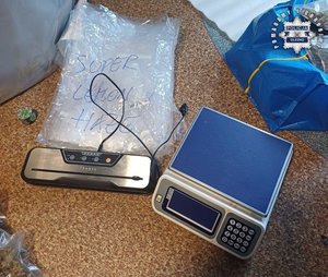 Zdjęcie przedstawia położone obok siebie na podłodze urządzenia : waga elektroniczna oraz zgrzewarka do folii. W prawym górnym rogu fragment niebieskiej torby z narkotykami.