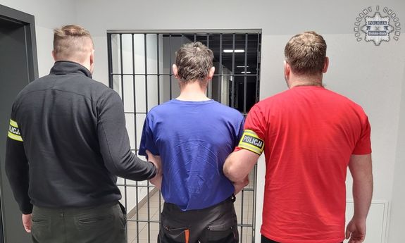 Dwaj ubrani po cywilnemu policjanci z zatrzymanym stoją przed kratą.