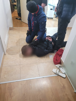 Zatrzymany mężczyzna leży na ziemi, przy nim kuca zatrzymujący go policjant. Obok stoi drugi policjant. Obaj funkcjonariusze są nieumundurowani.