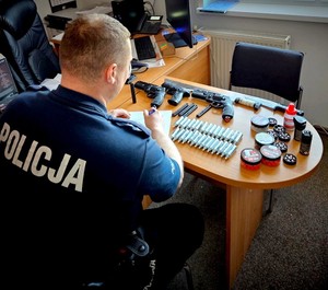 Policjant siedzi przy stoliku, na którym leżą zabezpieczone naboje i broń.
