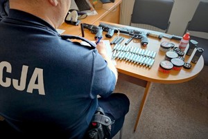Policjant siedzi przy stoliku, na którym leżą zabezpieczone naboje i broń.