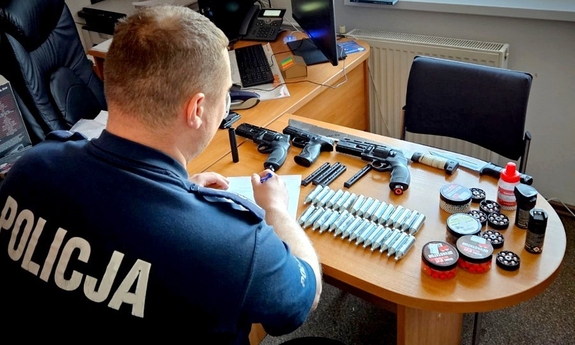 Policjant siedzi przy stoliku, na którym leżą zabezpieczone naboje i broń.