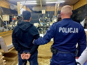 Policjant w mundurze trzymający za ramię mężczyznę z kajdankami założonymi na ręce trzymane z tyłu w tle maszyny do produkcji papierosów.