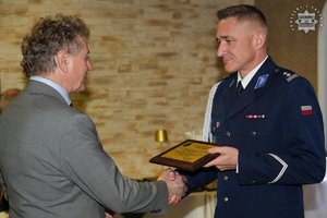 Umundurowany policjant odbiera z rąk mężczyzny w garniturze pamiątkową tabliczkę.