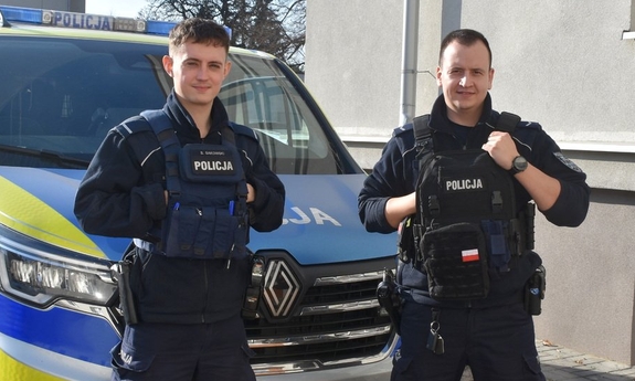 Dwaj policjanci stoją przed radiowozem policyjnym.