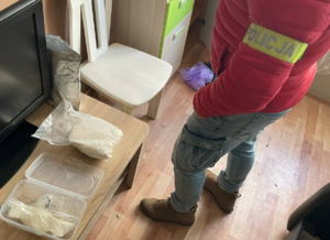 Zabezpieczone narkotyki w foliowym woreczku i plastikowym pudełku leżą na stoliku, obok stoi funkcjonariusz.