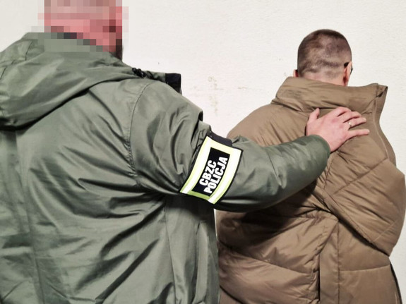 Funkcjonariusz CBZC, w opasce na ręce z napisem CBZC, trzyma dłoń na barku stojącego przed nim mężczyzny. Widok z boku.