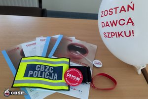 Na stoliku lezą ulotki, opaska z napisem CBZC Policja, przypinki DKMS i balon z napisem Zostań dawca szpiku!