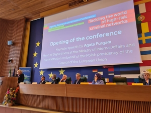 Uczestnicy konferencji siedzą przy stole. Na ścianie za nimi znajduje się ekran, na którym wyświetlana jest prezentacja. Po lewej stronie kobieta stoi przy mównicy przemawiając do zgromadzonych.