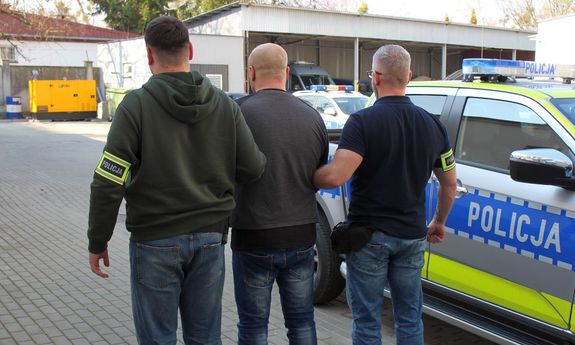 Dwaj ubrani po cywilnemu policjanci prowadzą zatrzymanego. Obok stoi zaparkowany policyjny radiowóz.