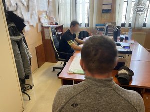 Na zdjęciu policjant z opaską na ramieniu przy biurku, przy którym siedzi mężczyzna tyłem do obiektywu.