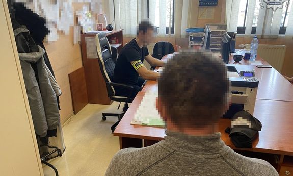 Na zdjęciu policjant z opaską na ramieniu przy biurku, przy którym siedzi mężczyzna tyłem do obiektywu.