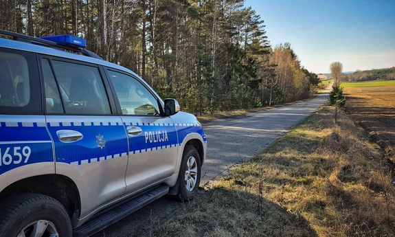 Policyjny radiowóz stoi na asfaltowej drodze biegnącej wzdłuż lasu.
