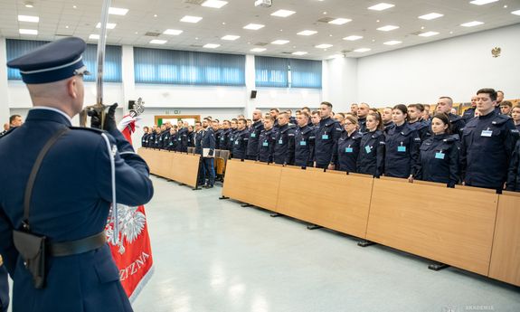 Z boku widoczny policjant z pocztu sztandarowego, który w dłoni trzyma uniesioną do góry szablę, a przed nim widoczni zebrani na sali słuchacze.