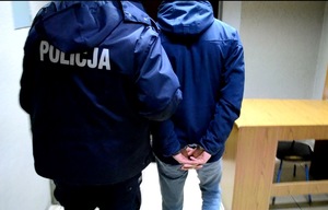Policjant w kurtce z napisem Policja na plecach z zatrzymanym, który na rękach trzymanych z tyłu ma założone kajdanki. Widok z tyłu.