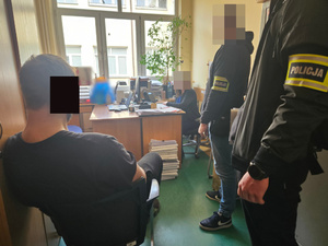 Zatrzymany mężczyzna siedzi na krześle podczas przesłuchania, obok stoją funkcjonariusze.