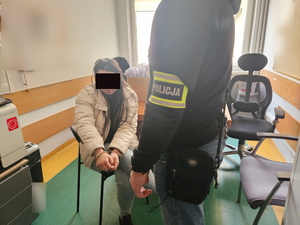 Zatrzymana kobieta w kajdankach siedzi na krześle, obok niej stoi funkcjonariusz.